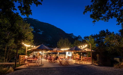 Café al aire libre con luces y montañas al fondo por la noche en Huttopia Gorges du Tarn, Occitania, Francia.