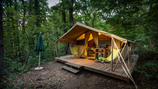 Tenda safari Bungalow tent Classic su piattaforma di legno nella foresta con sedie, tavolo e ombrellone.
