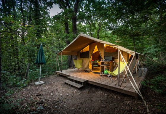 Safari tent Bungalow tent Classic on a wooden platform in the forest with chairs, table, and umbrella.