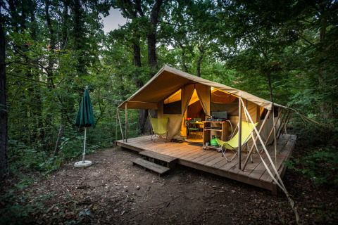 Tenda safari Bungalow tent Classic su piattaforma di legno nella foresta con sedie, tavolo e ombrellone.