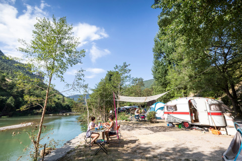 People camping by the river at Huttopia Gorges du Tarn in Occitanie, France, with caravan, tent, and trees.