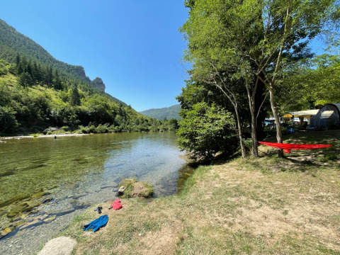 Scenic riverside camping site at Huttopia Gorges du Tarn, Occitanie, France, with forested hills and a hammock.