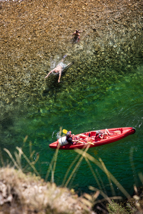Dos personas pasean en kayak rojo y dos nadan en agua cristalina junto a la orilla rocosa en Huttopia Gorges du Tarn.