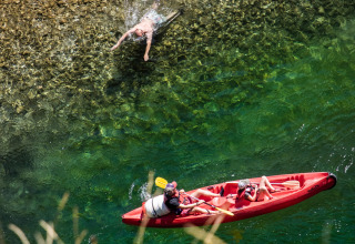 Two people paddle a red kayak, two others swim in clear water near a rocky shore at Huttopia Gorges du Tarn.