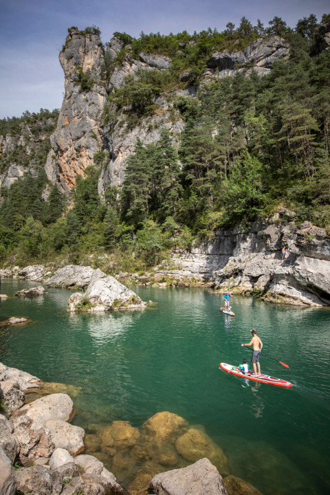People paddleboarding on a scenic river surrounded by cliffs and forest near Massegros-Causses-Gorges, France.