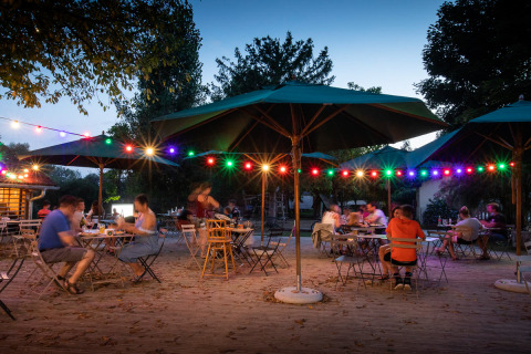 Cena al aire libre en Huttopia La Plage Blanche, Francia, con guirnaldas de luces y sombrillas al atardecer.