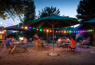 Cena all'aperto a Huttopia La Plage Blanche, Francia, con luci colorate e grandi ombrelloni al tramonto.