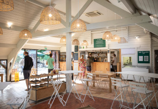 People waiting in a bright café with white furniture and wicker lamps at Huttopia La Plage Blanche, France.