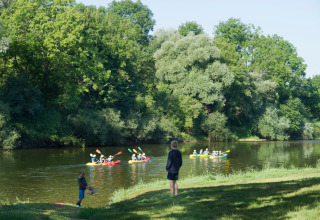 Children watch kayakers paddle on a calm river surrounded by lush green trees in Bourgogne-Franche-Comté, France.