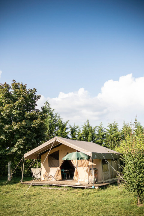 Spacious tent lodge with porch and chairs set amidst greenery at Huttopia La Plage Blanche holiday park, France.