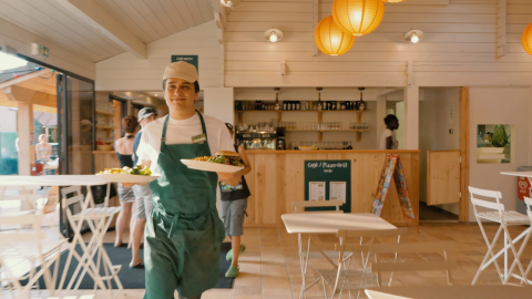 Un camarero con delantal verde sirve comida en un restaurante luminoso en Huttopia La Plage Blanche, Francia.