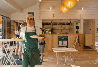 Een ober met een groene schort bedient borden in een gezellig restaurant in Huttopia La Plage Blanche, Frankrijk.