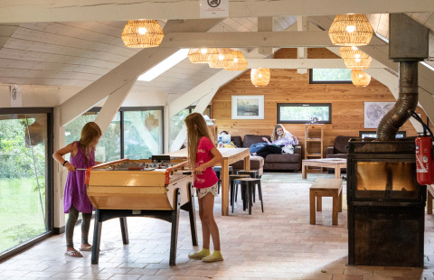 Two children play foosball in a cozy lodge with large windows, while an adult relaxes on the sofa behind them.