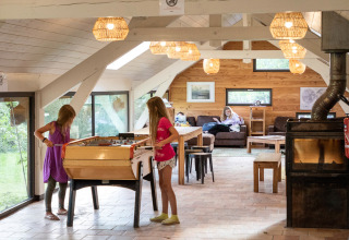 Zwei Kinder spielen Tischfußball in einer gemütlichen Hütte, während eine Frau auf dem Sofa entspannt.
