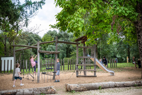 Niños juegan en un parque infantil de madera rodeado de árboles en Huttopia La Plage Blanche, Bourgogne-Franche-Comté.