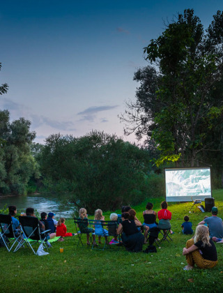 Familias disfrutan de una película al aire libre junto al río al atardecer en Huttopia La Plage Blanche, Francia.
