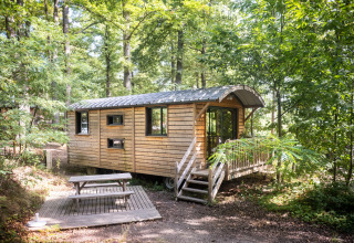 Houten hut omringd door bos bij vakantiepark Huttopia Wattwiller in Grand Est, Frankrijk, met picknickbank.