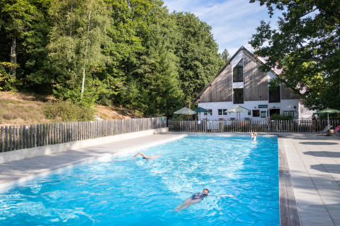 Piscina exterior en Huttopia Wattwiller, parque vacacional rodeado de bosque y terrazas con sombrillas.