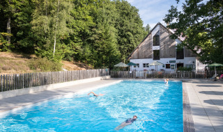 Piscina exterior en Huttopia Wattwiller, parque vacacional rodeado de bosque y terrazas con sombrillas.