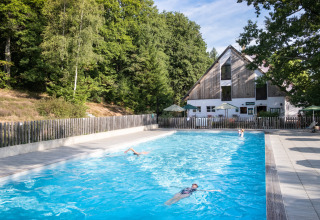 Udendørs swimmingpool ved Huttopia Wattwiller feriepark, omkranset af skov og terrasse med parasoller.