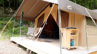 Safari telt Sweet på trædæk i skovmiljø, med stole og udendørs udstyr under overdækket terrasse.