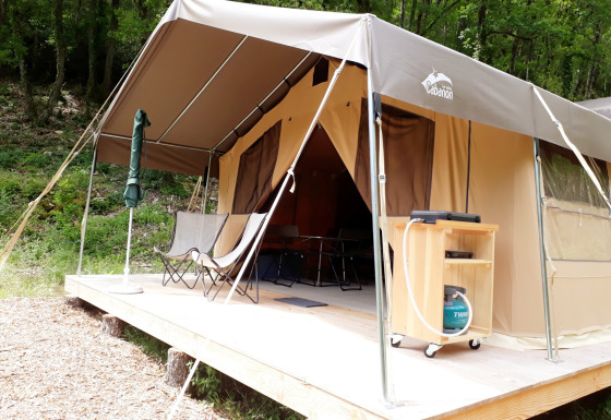 Tienda safari Sweet sobre plataforma de madera en el bosque, con sillas y equipo de camping exterior.
