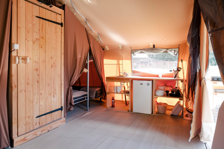 Interior of a safari tent featuring a wooden door, a kitchenette with fridge, shelves and a bunk bed.