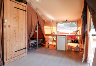 Interieur van een safaritent met houten deur, kitchenette met koelkast en stapelbed in de hoek.