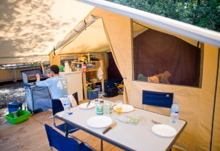 Inside a safari tent camp showing a dining table, kitchen area, and a person cooking at a stove.