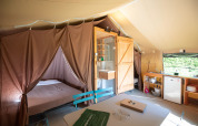 Interior view of a safari tent featuring a bed, bathroom, kitchenette, and dining table in natural light.