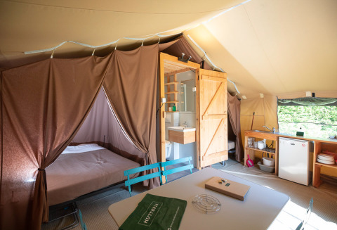 Interior view of a safari tent featuring a bed, bathroom, kitchenette, and dining table in natural light.