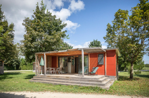 Casa mobile Vancouver con veranda in legno, sedie da giardino e alberi verdi sotto un cielo soleggiato.