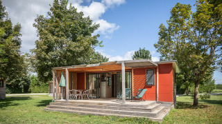 Mobile home Vancouver-lodge met houten veranda, tuinstoelen, bomen en gras onder een blauwe lucht.
