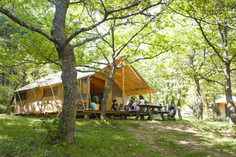 Family relaxing in front of a safari tent in a lush forest, surrounded by green trees and sunlight.