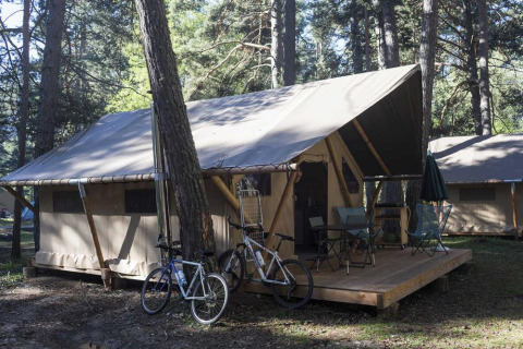 Safari telt Trappeur med træterrasse, to cykler og campingmøbler i skovområdet Huttopia Wattwiller.
