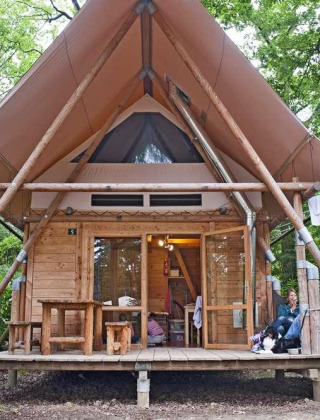 Een Cahutte safaritent in het bos met houten wanden, overdekte veranda en mensen die buiten relaxen.