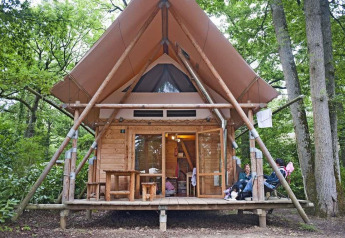A Cahutte safari tent in a forest setting, featuring wooden walls, a covered porch and people relaxing.