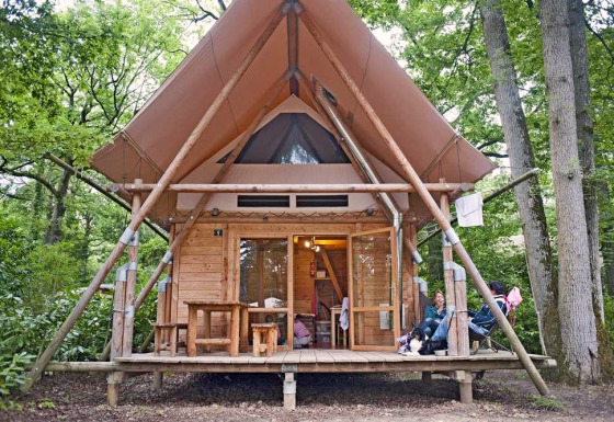 A Cahutte safari tent in a forest setting, featuring wooden walls, a covered porch and people relaxing.