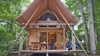 A Cahutte safari tent in a forest setting, featuring wooden walls, a covered porch and people relaxing.