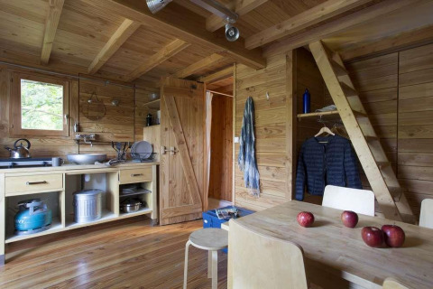Vista interior de una tienda safari Cahutte de madera con cocina, mesa comedor y escalera en Huttopia Wattwiller.