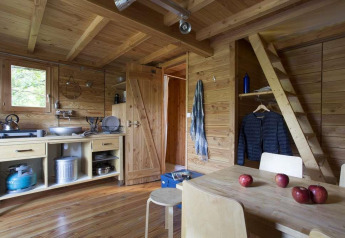 Vista interior de una tienda safari Cahutte de madera con cocina, mesa comedor y escalera en Huttopia Wattwiller.