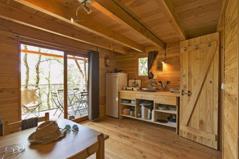 Acogedora cabaña de madera con cocina y comedor, puertas correderas a terraza con mesas y vista al bosque.
