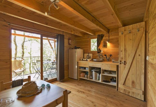 Acogedora cabaña de madera con cocina y comedor, puertas correderas a terraza con mesas y vista al bosque.