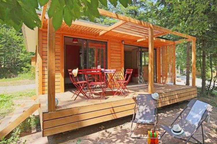 Cabaña Chalet Ottawa de madera con terraza, mesa y sillas rojas más tumbonas, rodeada de árboles.