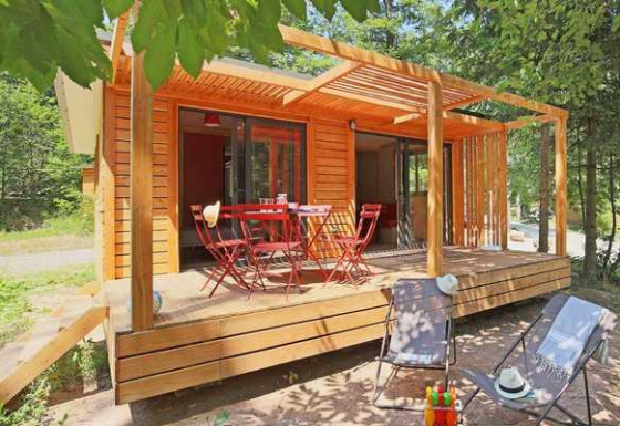 Cabaña Chalet Ottawa de madera con terraza, mesa y sillas rojas más tumbonas, rodeada de árboles.