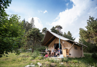 Famiglia che si rilassa davanti a una tenda safari Canadienne Tent II a Huttopia Lac de Serre Ponçon, tra gli alberi.