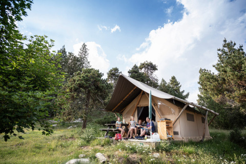 Familia relajándose frente a una tienda safari Canadienne Tent II en Huttopia Lac de Serre Ponçon, rodeados de árboles.