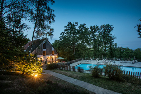 Vista nocturna de un albergue de madera iluminado y piscina rodeados de bosque en Huttopia Wattwiller, Francia.