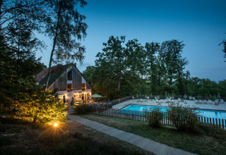 Avondzicht op een verlichte lodge en zwembad omringd door bos bij Huttopia Wattwiller in Frankrijk.