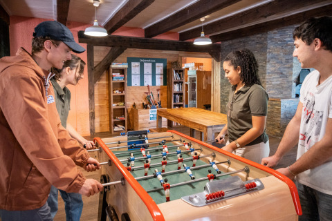 Cuatro jóvenes adultos juegan al futbolín en una sala común acogedora en Huttopia Wattwiller, parque turístico en Grand Est, Francia.